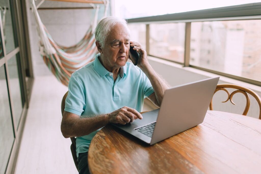 Free An Elderly Man Speaking Thought the Smartphone by the Laptop Stock Photo